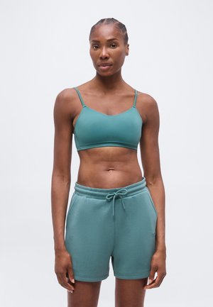 Nő, aki türkiz sportmelltartót és hozzáillő húzózsinóros rövidnadrágot visel, egyszerű fehér háttér előtt áll.