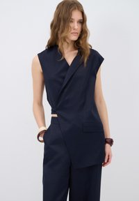 Marinblå ärmlös topp med omslagdesign, försedd med rak krage, sidoknutdetalj och textil med struktur; matchas med tillhörande byxor.