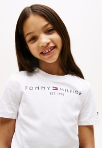 Bela bombažna majica s kratkimi rokavi, na kateri je napis "TOMMY HILFIGER EST. 1985" v temno modri barvi. Vključuje majhen logotip na rokavu in klasičen krožni izrez.