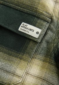 Detail zeleno-beige kockovanej tkaniny s bielou etiketou, na ktorej je vyšité "HRÁME BEZ LIMITOV Od osemdesiatdva".