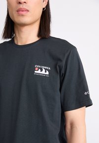 Zwart T-shirt met korte mouwen van katoen, met een logo op de linkerborst en een ander logo op de rechtermouw. Eenvoudig en strakke ontwerp.