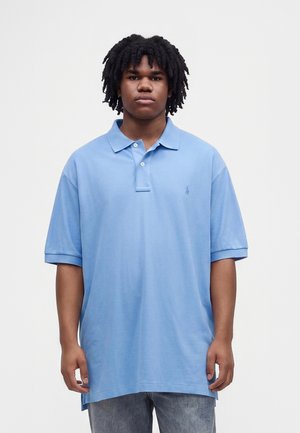 Jeune homme à la peau métisse et aux cheveux noirs bouclés porte un polo bleu clair ample et un jean gris sur un fond blanc uni.