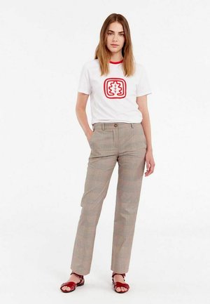 T-shirt blanc avec col rouge et motif graphique, associé à un pantalon à carreaux légers. Le modèle porte des sandales rouges et se tient devant un fond blanc.