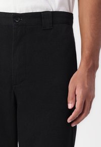 Black trousers made from a textured fabric, featuring a button closure, belt loops, and side pockets. Close-up of waistband and hand.