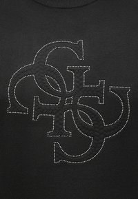 Melnā džemperī ar teksturētu dizainu, kurā pārklājas burti, izcelti ar maziem metāla kniedēm, demonstrējot rakstu kontrastu.