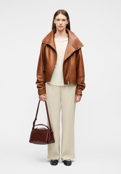 Γυναίκα που φοράει καφέ φαρδιά δερμάτινη ζακέτα, λευκό πουκάμισο, μπεζ παντελόνι, μαύρα παπούτσια, κρατώντας μια σκουρόκαφέ τσάντα με μακρύ ιμάντα.