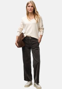 Maglione beige a scollo a V, pantaloni in corduroy marrone scuro a gamba larga e sneakers chiare. La modella tiene una borsa in suede marrone.
