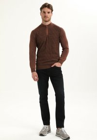 Brązowy sweter z dzianiny z kołnierzem na pół-zamek i teksturowanym geometrycznym wzorem, w połączeniu z ciemnymi dżinsami i szarymi butami sportowymi.