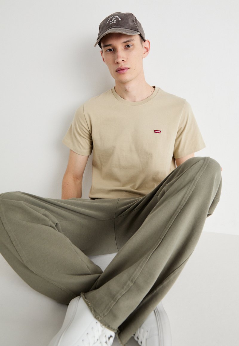 Camiseta de algodón beige con un pequeño logo rojo, pantalones verdes oliva con detalles en las costuras y zapatillas blancas. Persona llevando una gorra gris.