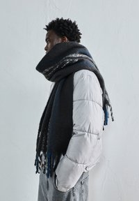 Bărbat îmbrăcat cu o eșarfă mare, de culoare albastru închis și gri, și o geacă puffer de un gri deschis, stând deoparte cu mâinile în buzunare.