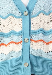 Lyseblå cardigan med dekorative strikkede mønstre i hvite og oransje striper, med seks knapper og ribbet bunnkant.
