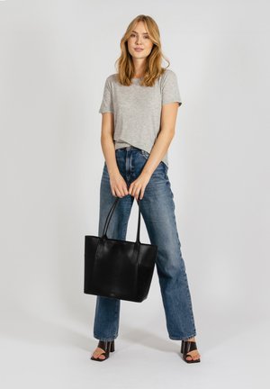 Black leather tote bag with two sturdy handles, rectangular shape, smooth texture. Worn with a grey t-shirt and blue denim jeans.