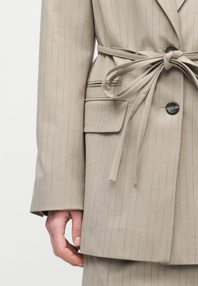 Μπεζ σακάκι με ρίγες, ζώνη φτιαγμένη από ύφασμα δεμένη στη μέση, τσέπη με καπάκι, μαύρα κουμπιά και ορατό αριστερό χέρι που ακουμπάει κοντά στο πλευρό.