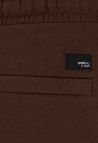 Bruine stof met een zachte textuur, een elastische tailleband, een zijzak en een zwart label met de tekst "spitzblub studios."