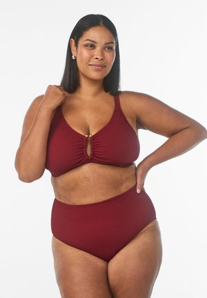 Pewna siebie kobieta w bordowym, teksturowanym kostiumie kąpielowym dwuczęściowym, z ręką spoczywającą na biodrze i ramieniu, stojąca na tle jednolitego białego tła.