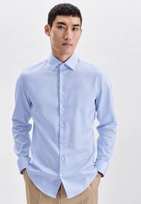 Lichtblauw overhemd met knopen, gemaakt van gladde stof met een klassieke kraag en lange mouwen. Heeft een rechte zoom en witte knopen.