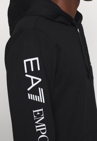 Sudadera con capucha negra con el texto blanco "EA" y "EMPORIO ARMANI" en la manga. Cuenta con cordones y una tela suave y texturizada.