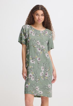 Kvinde med krøllet hår iført en grøn knælængde kortærmet kjole med pink og hvide blomsterprint, stående foran en ensfarvet baggrund.