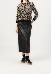 Kobieta w swetrze w panterkę, czarnej spódnicy midi z ekoskóry, czarnych skarpetkach i czarnych trampkach Adidas z białymi paskami.