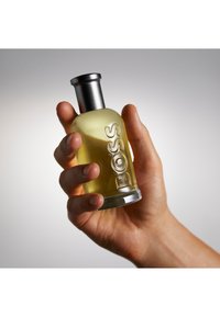 Hand die een cologneflesje vasthoudt met een gele tint, een zilveren dop en het woord "BOSS" in reliëf op de voorkant.