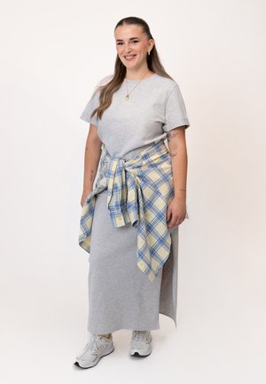 Frau mit langen Haaren, die ein graues Kleid, weiße Sneakers und ein gelb-blau kariertes Hemd um die Taille gebunden trägt, lächelt in die Kamera.