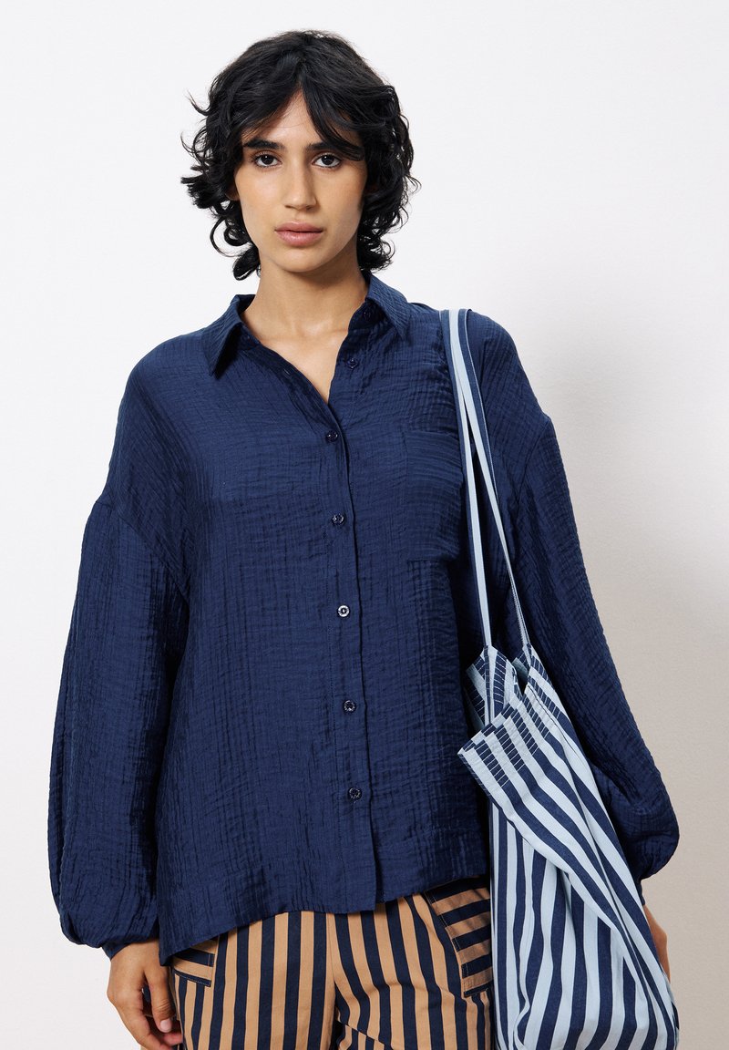 Chemise boutonnée texturée bleu marine avec des manches larges, associée à un pantalon rayé marron et bleu marine et un sac fourre-tout rayé.