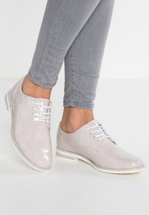 Jasnoszare skórzane buty o błyszczącym wykończeniu, z białymi sznurówkami, niskim obcasem i teksturowaną podeszwą. Nosi się je z dopasowanymi szarymi spodniami.