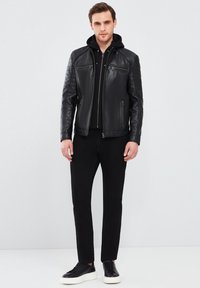 Chaqueta de cuero negra con acentos acolchados en los hombros, bolsillos con cremallera y un diseño ajustado, llevada sobre una sudadera negra con capucha y pantalones negros.