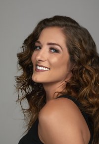 Portrait d'une femme avec des cheveux bruns mi-longs et bouclés. Elle a des yeux bleus, des lèvres brun clair et porte un débardeur noir. Fond neutre.