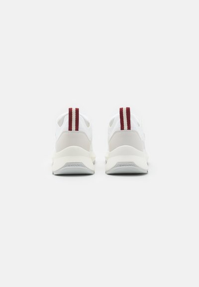 White sneakers featuring a smooth upper, red vertical stripes on the back, and a cushioned sole with the brand name embossed.