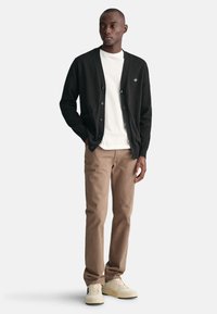Schwarze Strickjacke aus Strickmaterial mit V-Ausschnitt, Knopfleiste und zwei Fronttaschen. Getragen über einem weißen T-Shirt mit beigen Hosen.
