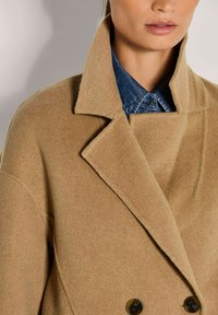 Vrouw die een camel-kleurige wollen jas draagt over een blauw denim overhemden met knopen, met een close-up van de kraag en knopen.