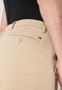 Pantaloni khaki in cotone con una texture liscia, caratterizzati da una tasca posteriore con bottone e un piccolo logo del brand in rosso, bianco e blu.