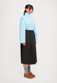 Sieviete gaiši zilā augsta apkakles blūzē, melnā midi svārku garumā, pelēkās zeķēs un brūnās platformas loafers, stāvot pret vienkāršu baltu fonu.
