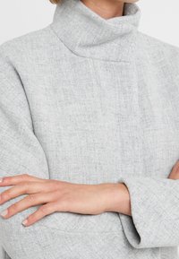 Personne portant un manteau à col roulé texturé gris clair, bras croisés, montrée du cou au milieu du torse, sur un fond uni.