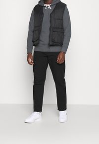 Gilet imbottito nero sopra una felpa grigia, abbinato a pantaloni neri e sneakers bianche. Il gilet ha quattro tasche anteriori e una chiusura con zip.