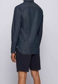 Marinblå långärmad skjorta med texturerad tyg, med rundad nederkant och knäppte manschetter, i kombination med mörka shorts.