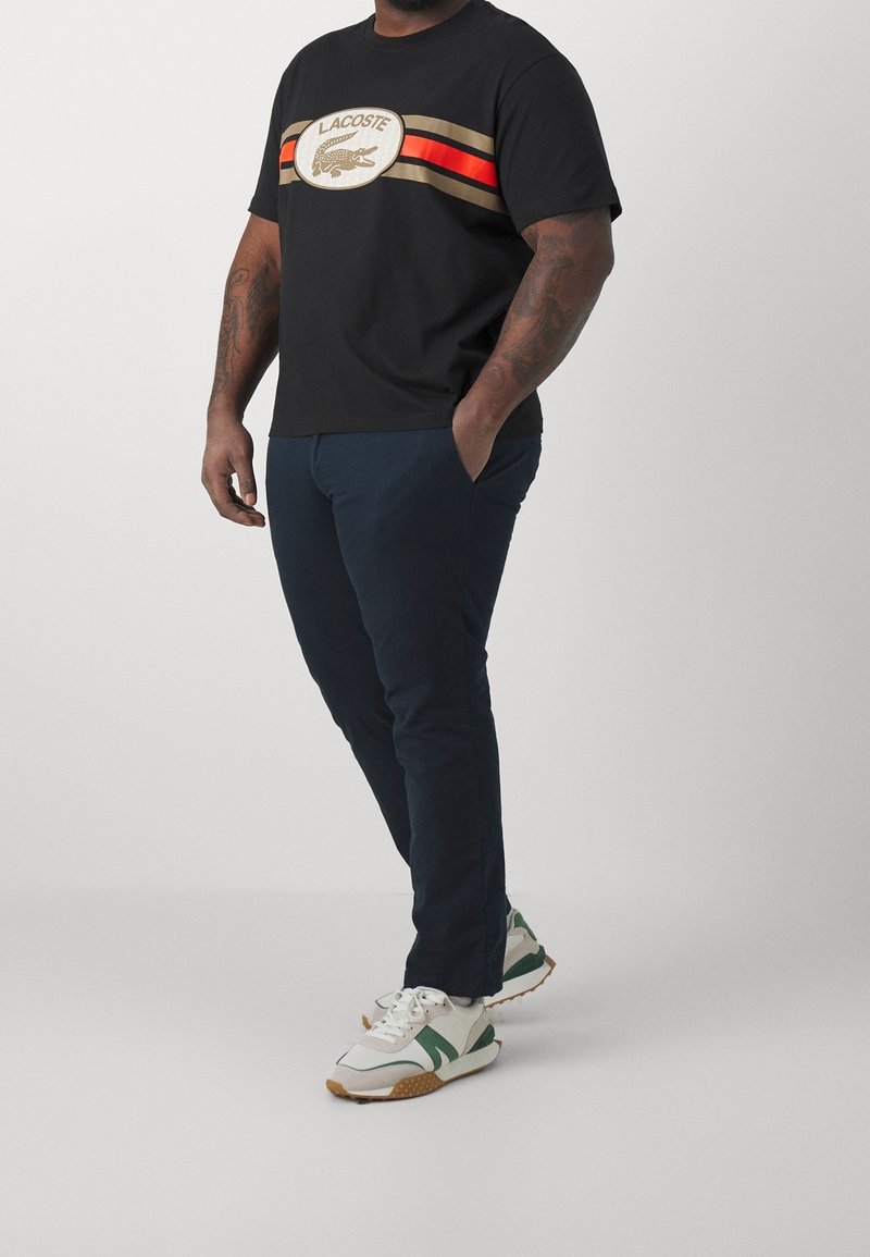 Sort t-shirt med Lacoste-logo og vandrette striber i guld, orange og beige. Kombineret med marineblå bukser og hvide-grønne sneakers.