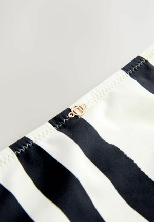 Mustvalge triibuline kangas, millel on sile tekstuur. Õmbluse lähedal on väike kuldse tooniga logo detail. Nähtav rõhutatud õmblus.