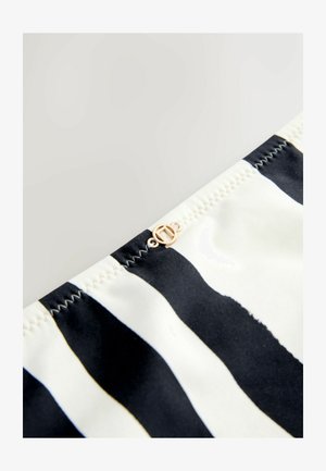 Mustvalge triibuline kangas, millel on sile tekstuur. Õmbluse lähedal on väike kuldse tooniga logo detail. Nähtav rõhutatud õmblus.