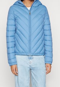 Světle modrá prošívaná péřová bunda s kapucí, s chevronovým prošívaným vzorem, zapínáním na zip a žebrovanými manžetami. Nošena přes světle modré džínové kalhoty.