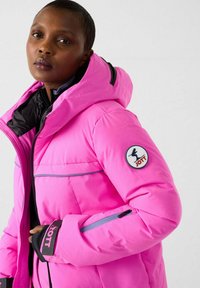 Donna che indossa una giacca a vento imbottita di colore rosa brillante con cappuccio, strato interno nero e toppa con il logo JOTT sulla manica sinistra, su sfondo chiaro uniforme.