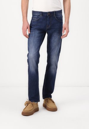 Homme portant un jean bleu foncé délavé et des chaussures en daim beige, debout sur fond blanc.