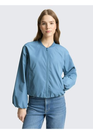 Jeune femme portant une veste bomber bleu clair zippée et un jean bleu, debout avec une main dans la poche sur un fond uni.