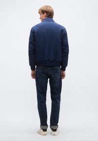 Marineblauwe bomberjack met een geribde kraag en manchetten, met een gladde buitenkant en een comfortabele pasvorm, gecombineerd met donkere spijkerbroek en sneakers.