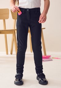 Musta slim-fit-housut nappilulla ja taskuilla, yhdistettynä kiiltäviin mustiin kenkiin. Kirkkaan pinkkejä kyniä näkyy sivutaskussa.