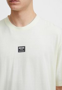 Lys gul bomulds t-shirt med korte ærmer, der har et kvadratisk sort mærke med teksten "INDICODE EST. 1993" og "CARRY OUR PASSION."