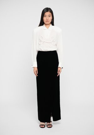 Witte blouse met opvallende lussen aan de voorkant, gecombineerd met een zwarte fluwelen rok met een zijsplit en zwarte sandalen.