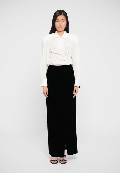 Camicia bianca con dettagli a cerchio prominenti sul davanti, abbinata a una gonna nera in velluto con uno spacco laterale e sandali neri.