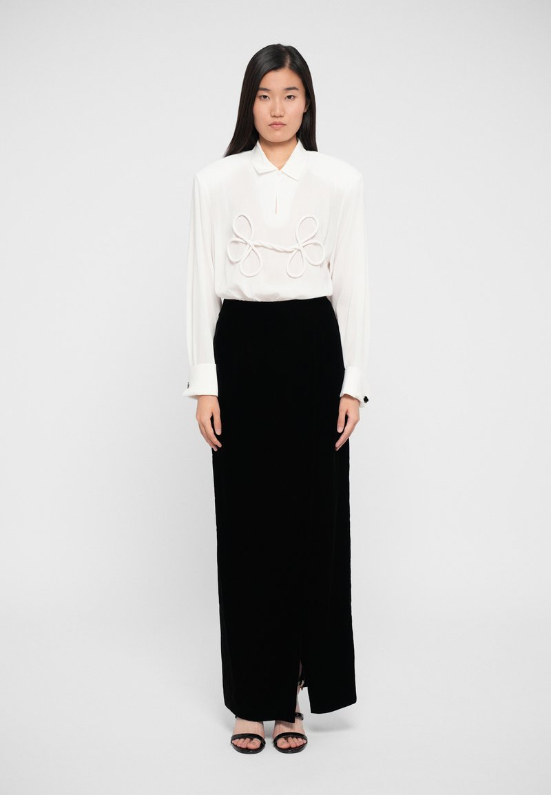 White blouse with prominent loop details at the front, paired with a black velvet skirt featuring a side slit and black sandals.